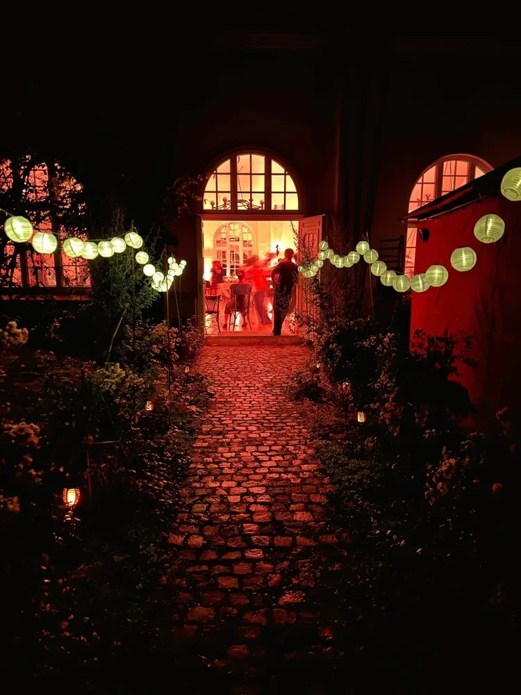 Ein dunkler Gartenweg mit Laternen führt zu einem hell erleuchteten Haus mit großen Fenstern. Im Inneren sind Menschen bei einer Feier zu sehen.