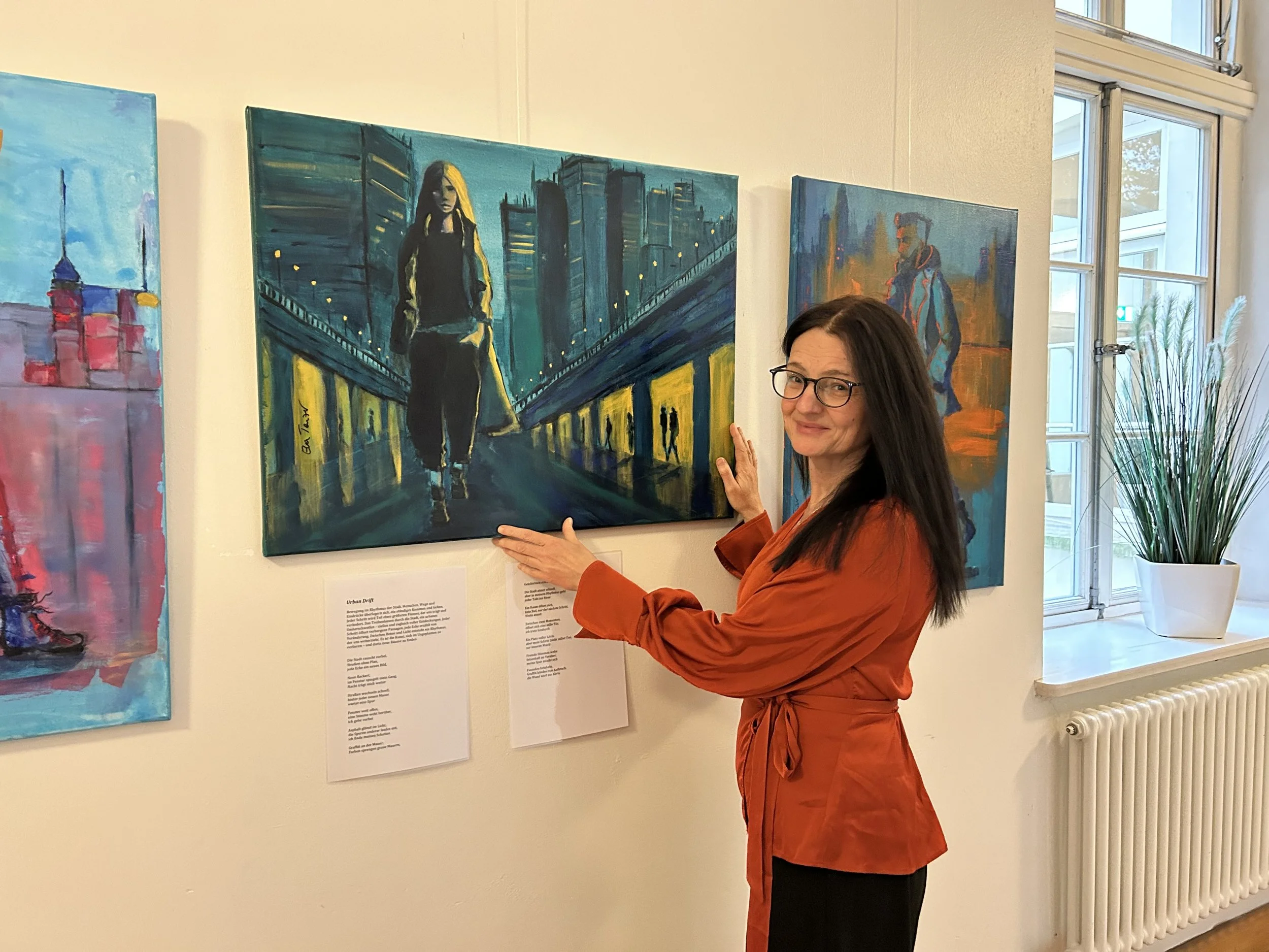 Frau in orange Jacke in Kunstgalerie neben Gemälde mit Stadtlandschaft und Person in der Mitte, zeigt auf das Gemälde, neben Fenster mit Vase und grünem Pflanzen