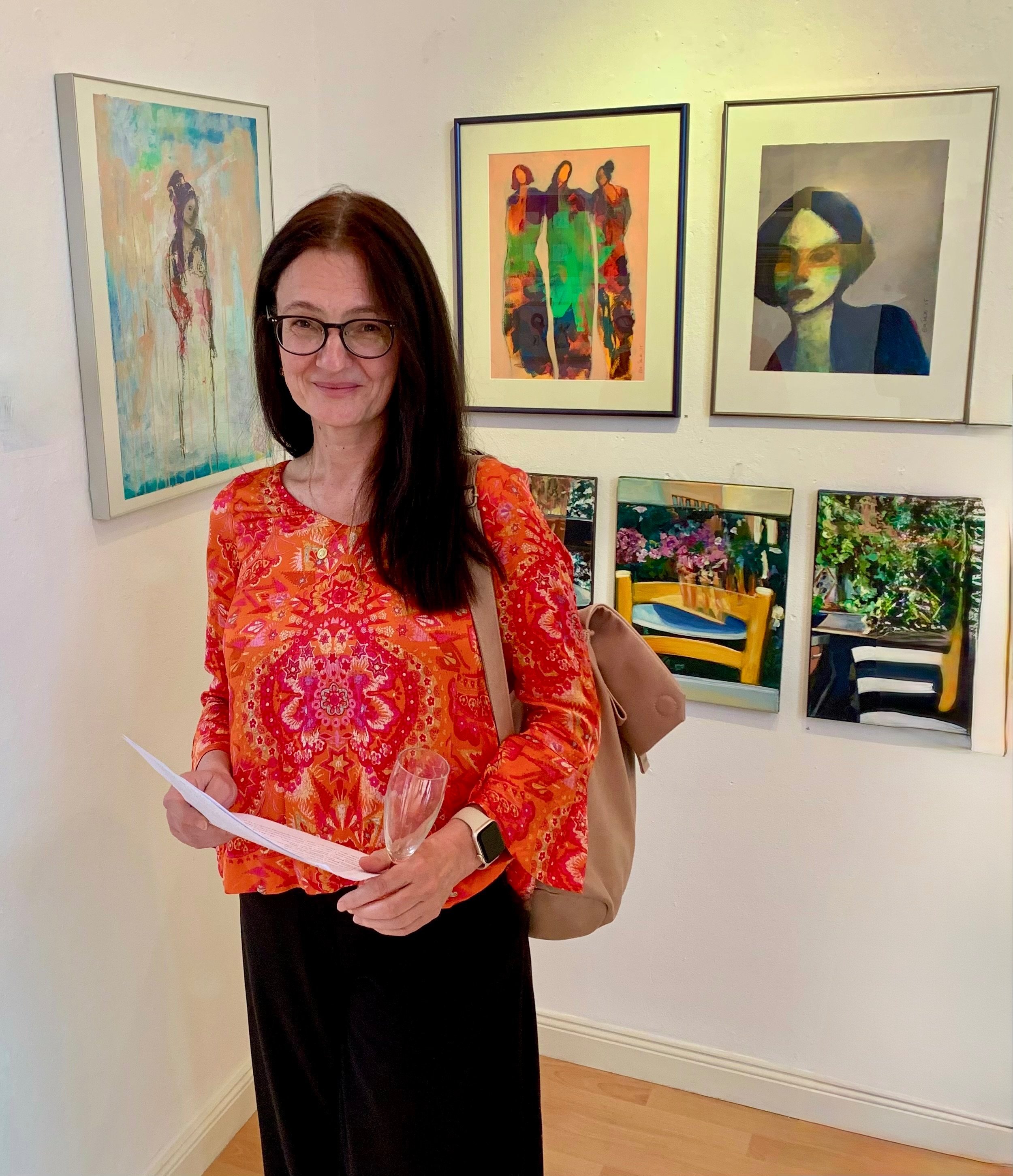 Eine Frau mit Brille, dunklem Haar und einem orangefarbenen, gemusterten Oberteil steht in einer Galerie, umgeben von Kunstwerken an der Wand.