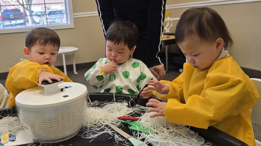 Three young children wearing yellow and patterned smocks working with tangled string and scissors at a table in a classroom.