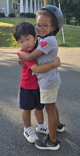 Two young boys hugging outdoors, one wearing a helmet and glasses, smiling, and the other with dark hair, wearing a red shirt and shorts.