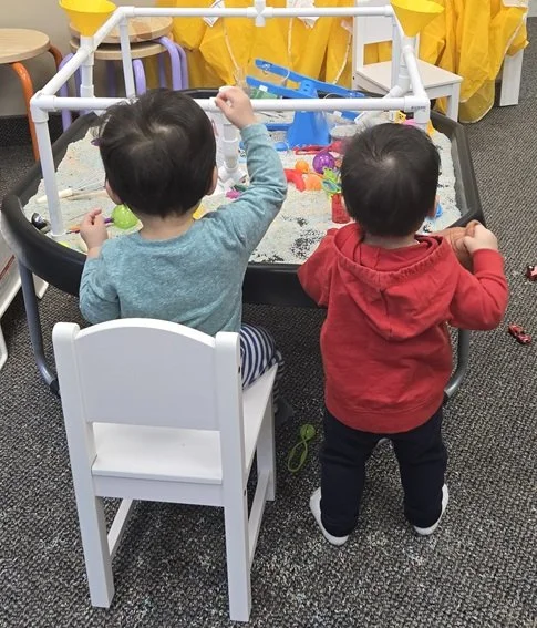 Two young children playing at a sensory table in an indoor play area. One child is sitting on a chair facing the table, while the other is standing beside it. Various toys and colorful items are on the table.