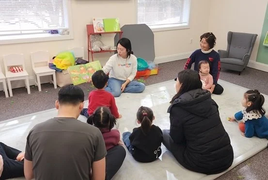 Adults and children sitting on a white mat in a room with windows, cushions, and chairs, participating in an activity or lesson.