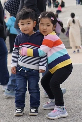 Two young children, a boy and a girl, hugging each other outdoors on a staircase with other people in the background.