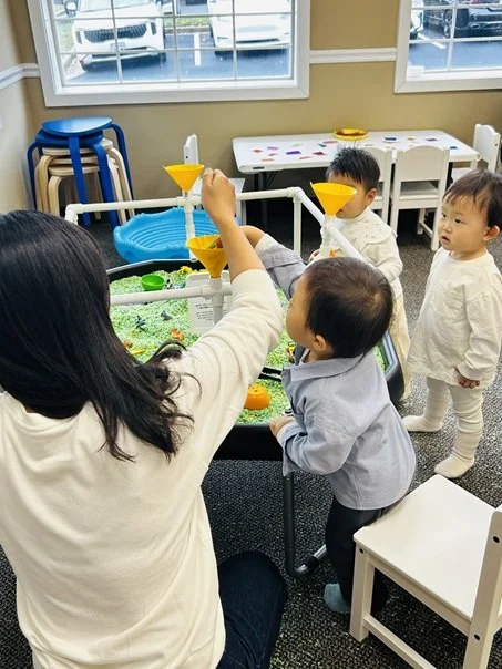 Children playing a game with a woman using a water or sand table in a classroom.