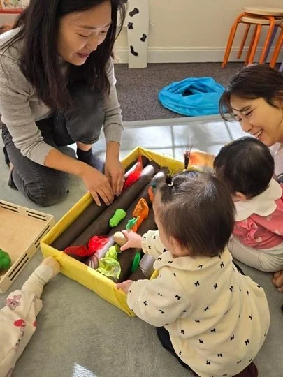 Two adults and two children playing with a toy set that looks like a gardening or outdoor play kit on the floor in a room.