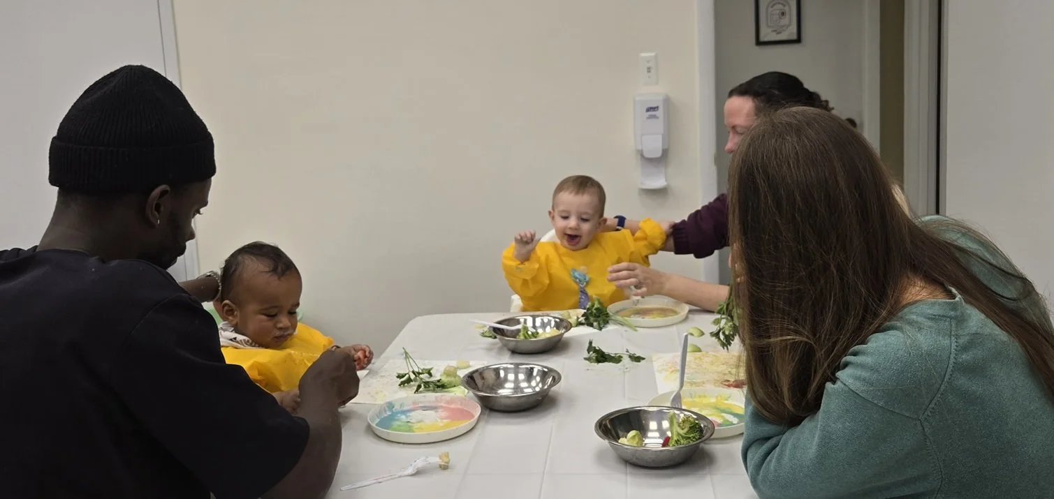 A group of four people, including two children and two adults, are seated at a dining table with bowls and plates. The children are wearing yellow bibs, and the table has remnants of food, salad greens, and utensils.