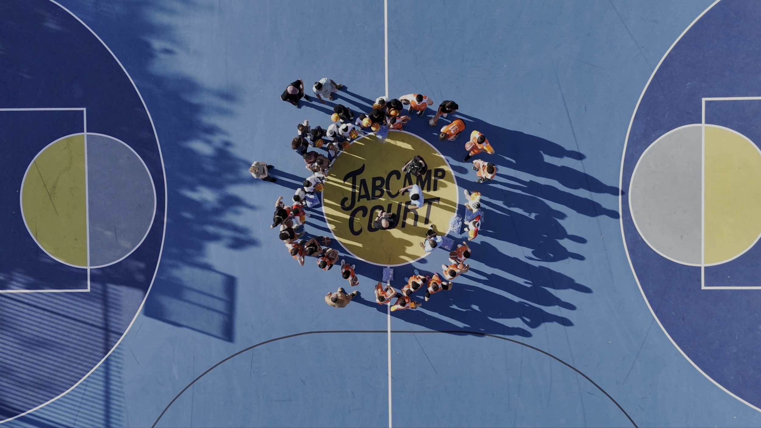 Overhead view of a group gathered in a circle at center court around the JAB Camp Court logo on a blue outdoor basketball court