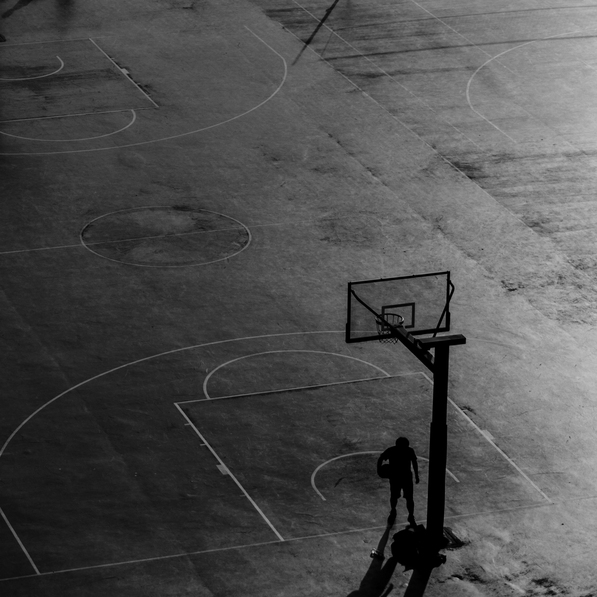 A solitary basketball player standing on an outdoor basketball court at dusk, casting a long shadow, with the hoop and backboard nearby.