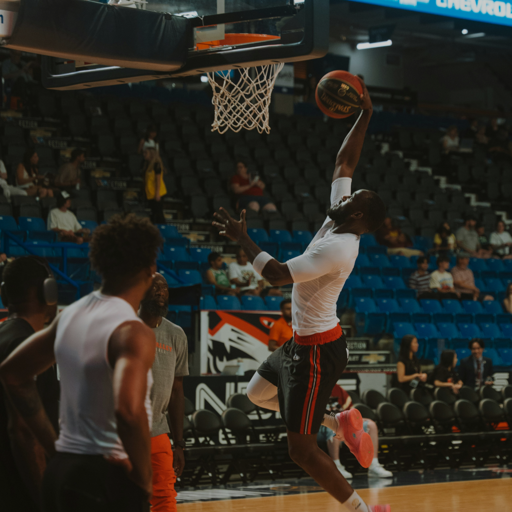A basketball player in a white shirt and black shorts is jumping near the hoop, about to dunk or lay up, with the ball in his right hand. Several onlookers and teammates are nearby on the court, and spectators are seated in the background.