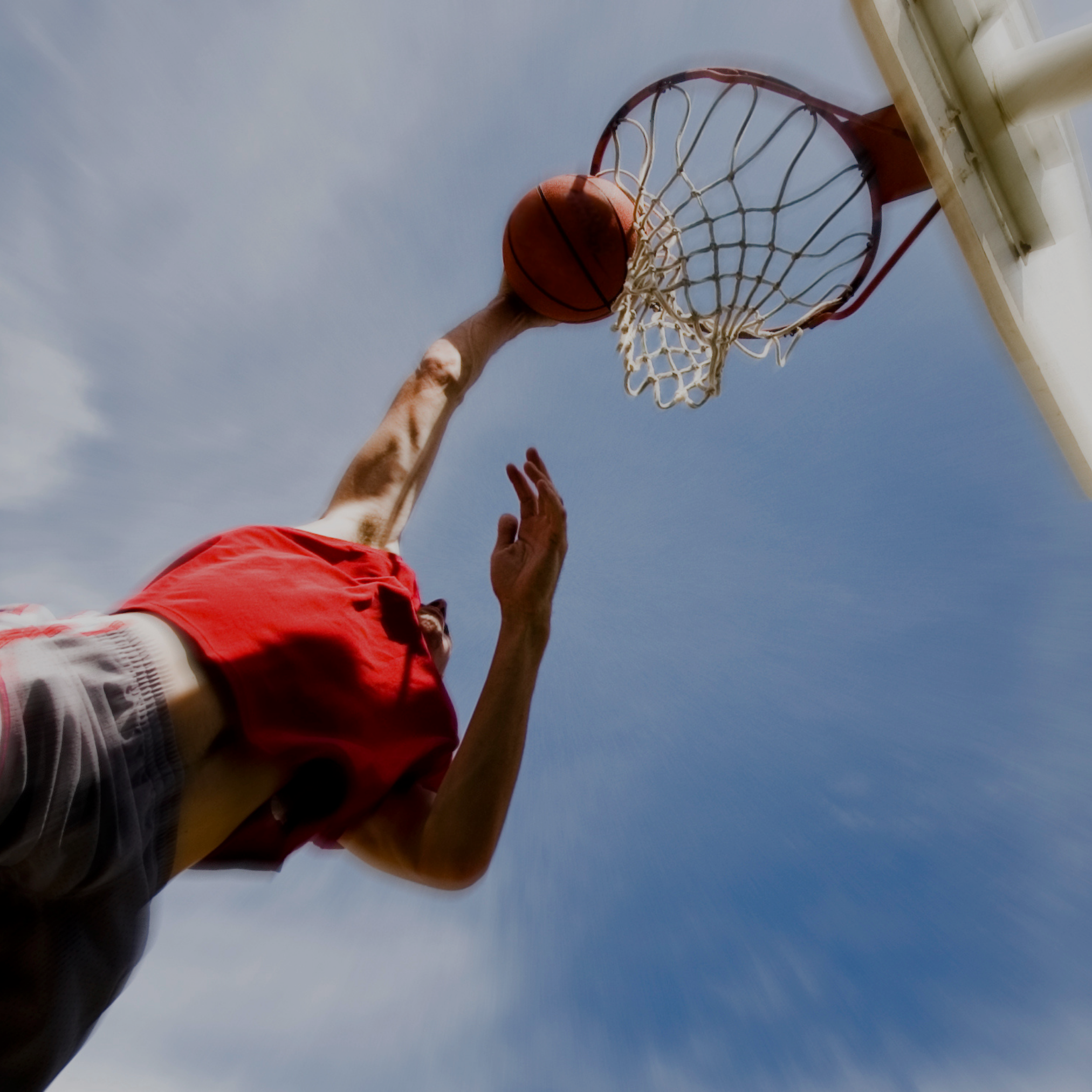 A person in a red shirt and shorts is playing basketball outdoors, about to dunk the ball into the hoop.