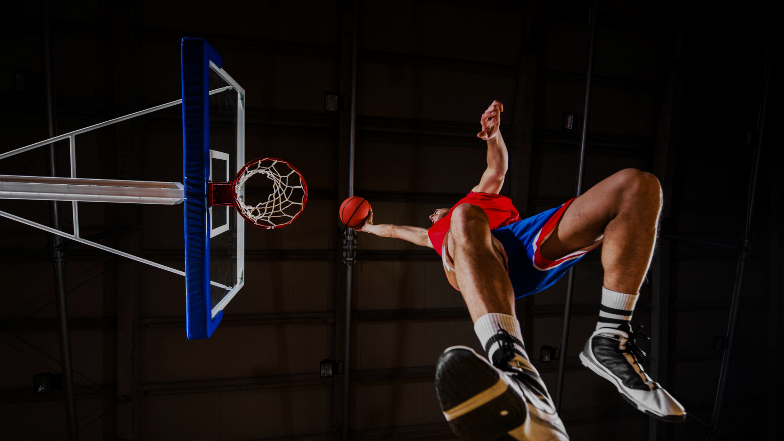 Low angle action shot of a basketball player mid dunk reaching toward the hoop inside an indoor gymnasium