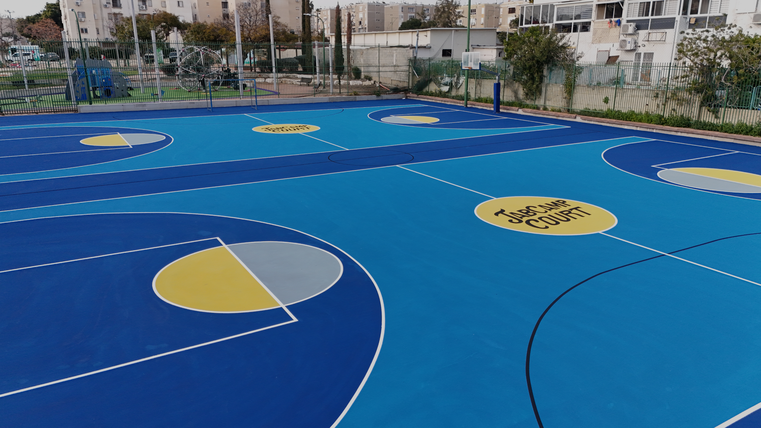 Empty outdoor basketball court with blue and yellow painted surface and JAB Camp Court logo at midcourt