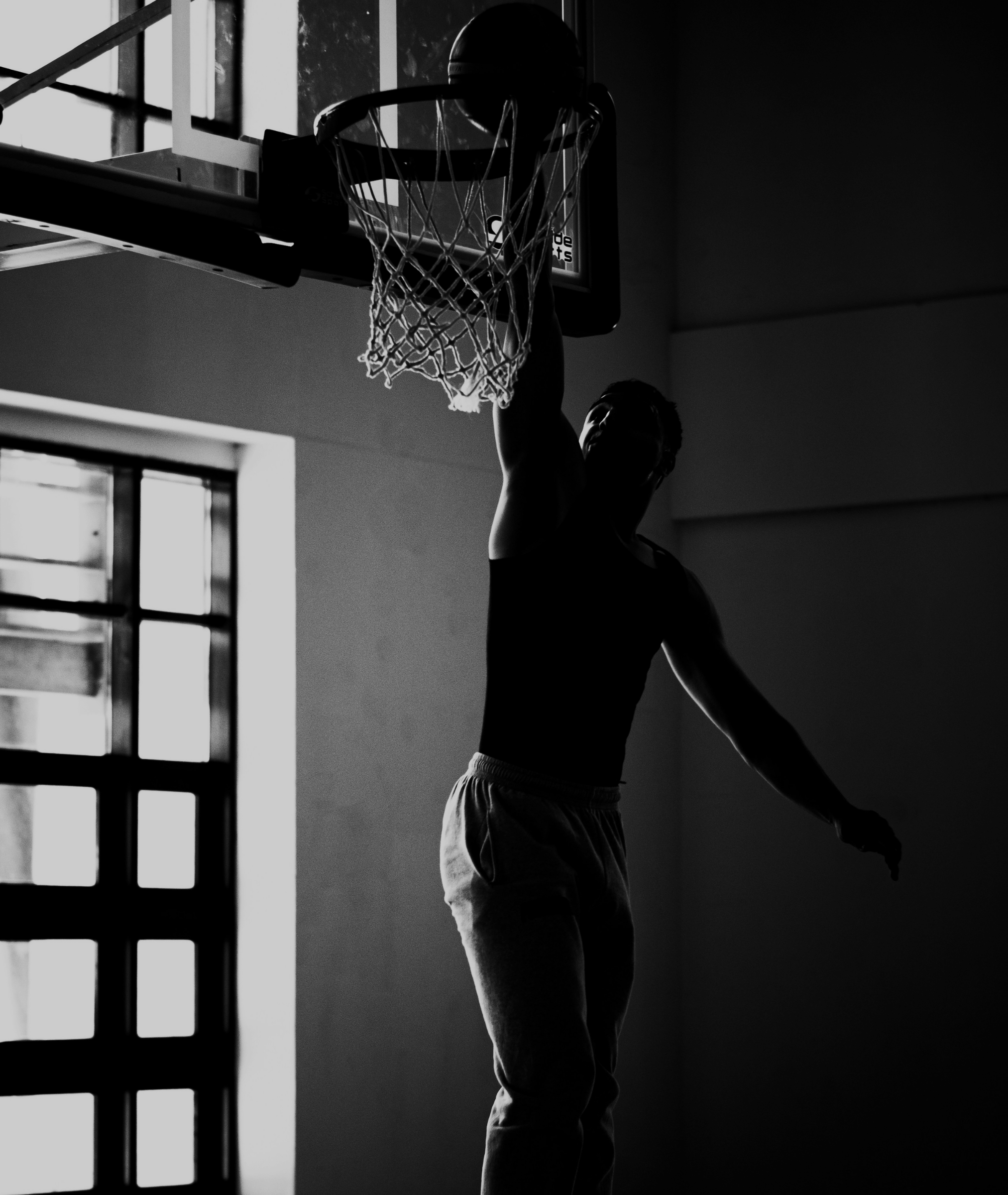 Silhouette of a basketball player dunking indoors near large windows with natural light streaming in