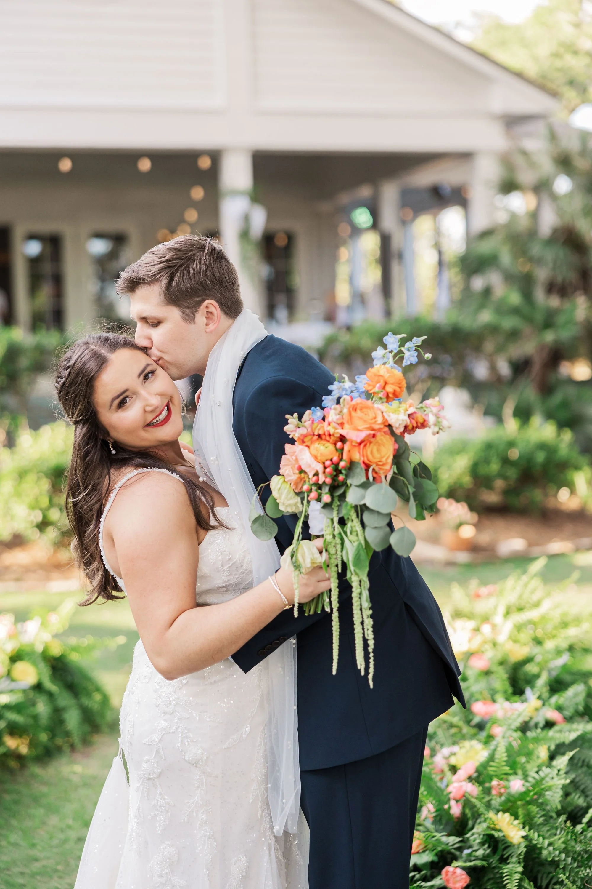bride-holding-colorful-bouquet-while-groom-kisses-her-forehe.jpg