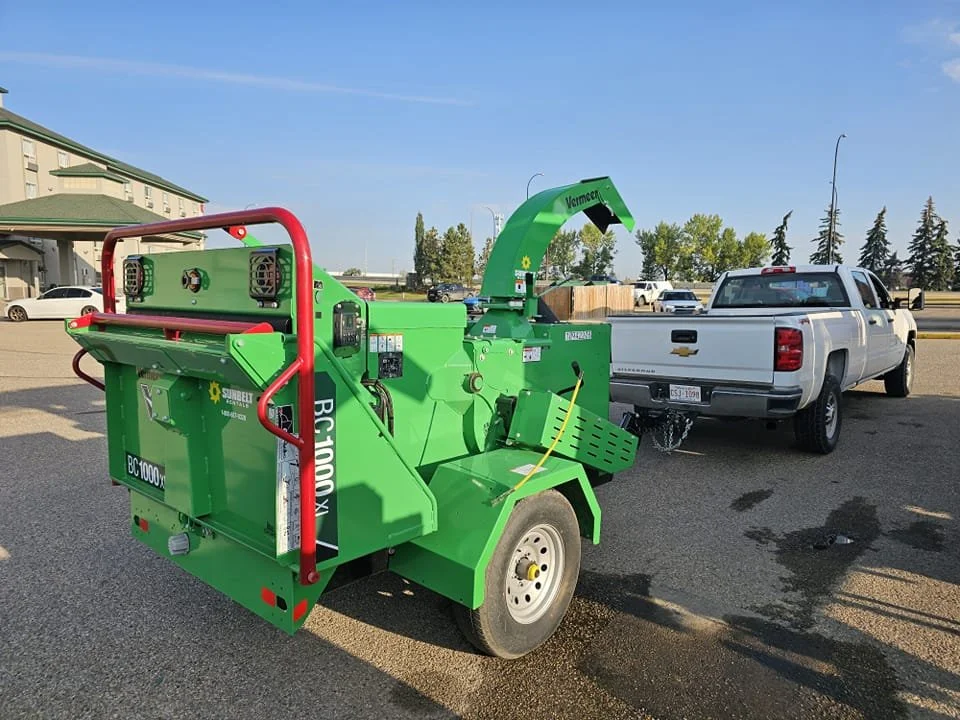 A green wood chipper attached to a white pickup truck in a parking lot on a sunny day.
