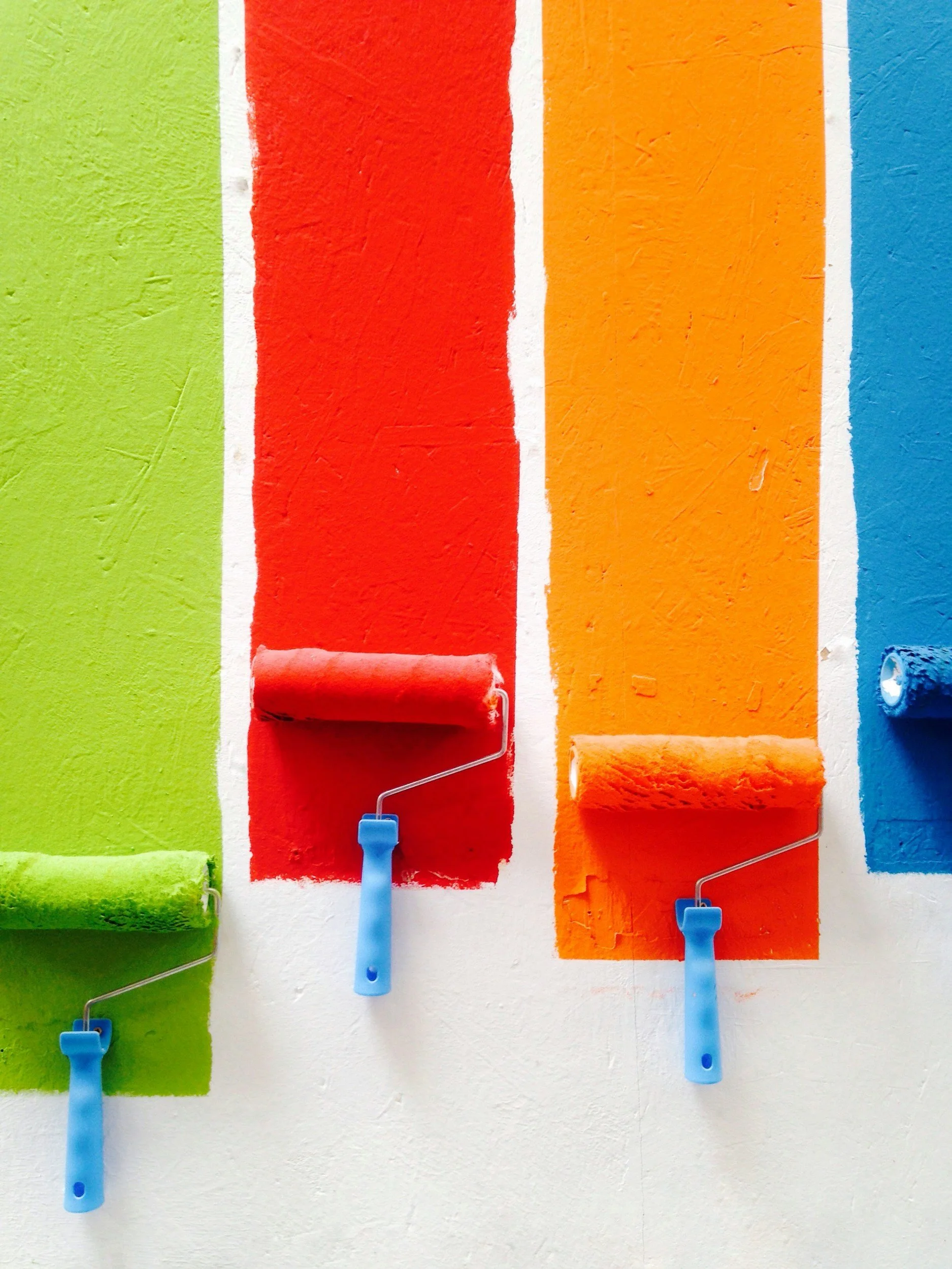Four paint rollers applying paint on a wall, each holding a different color: green, red, orange, and blue.