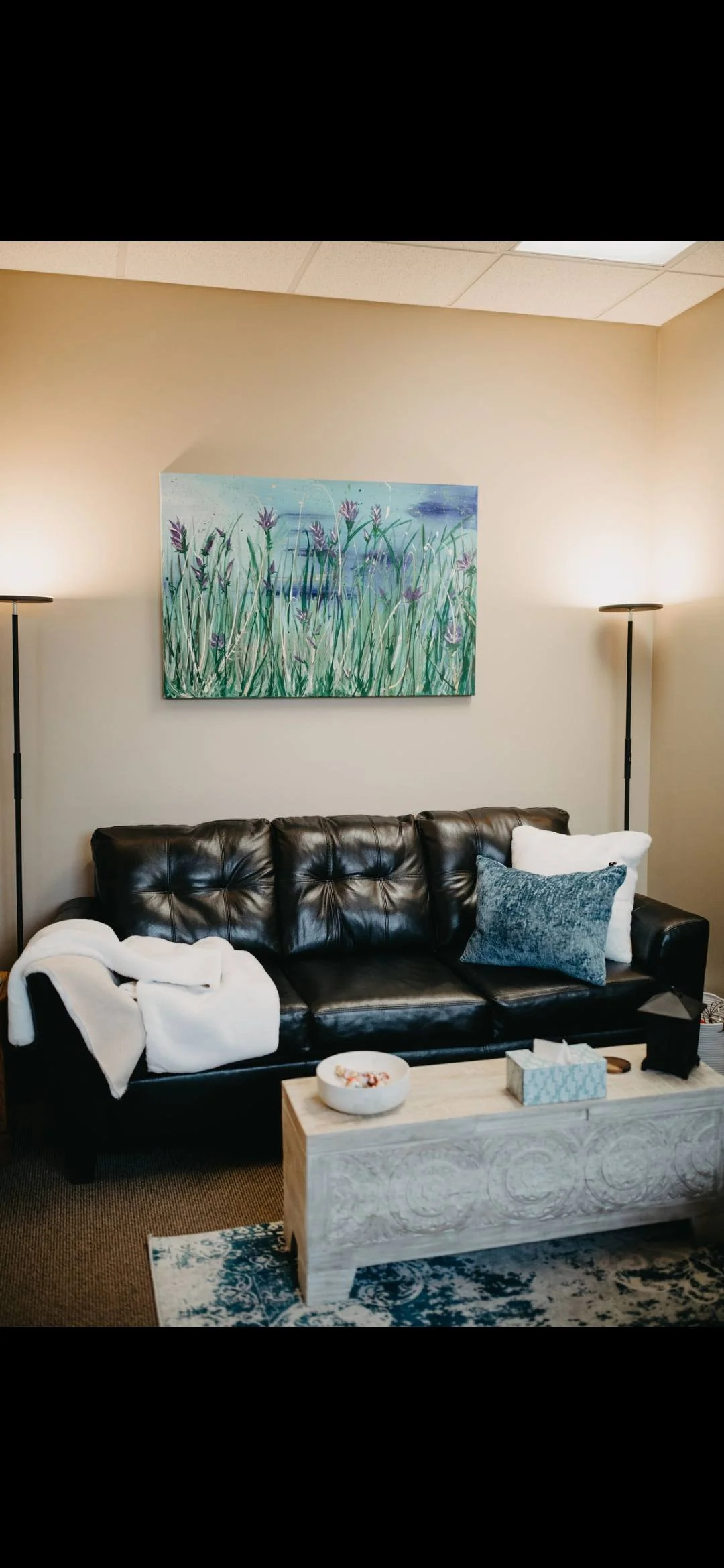 Living room with black leather sofa, white throw blanket, and cushions, placed in front of a beige wall with a colorful field of lavender painting. Two floor lamps stand on each side of the sofa, and a light wooden coffee table with decorative details holds a tissue box and a bowl of snacks. A patterned area rug covers part of the floor.
