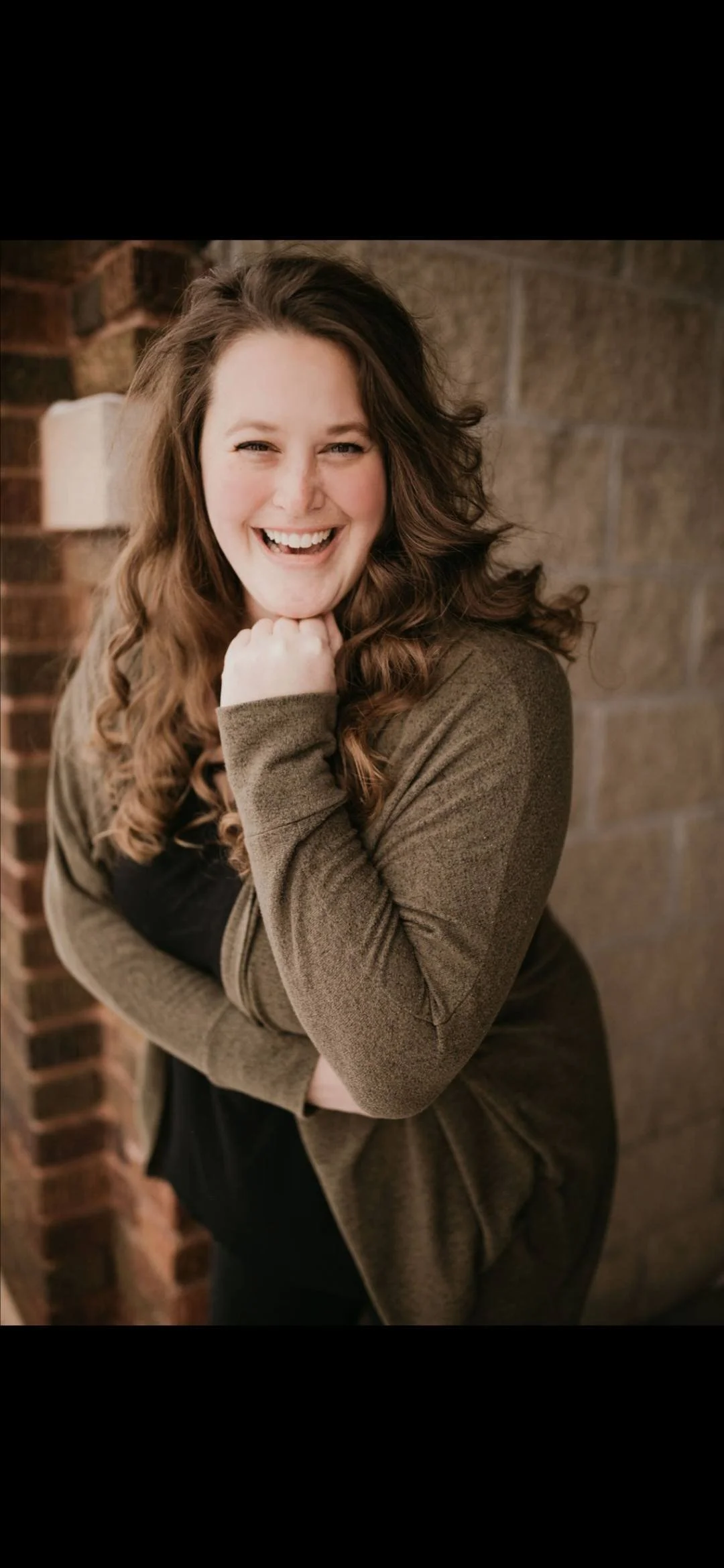 A woman with long, curly brown hair smiling and posing with her chin resting on her fist, leaning against a brick wall.