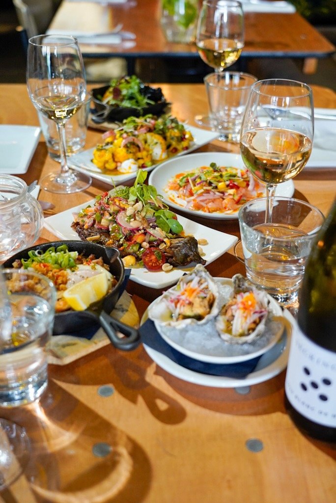 A wooden table set with several gourmet dishes, including oysters, grilled meat, colorful salads, and roasted vegetables, accompanied by glasses of white wine and water. A wine bottle is also visible.