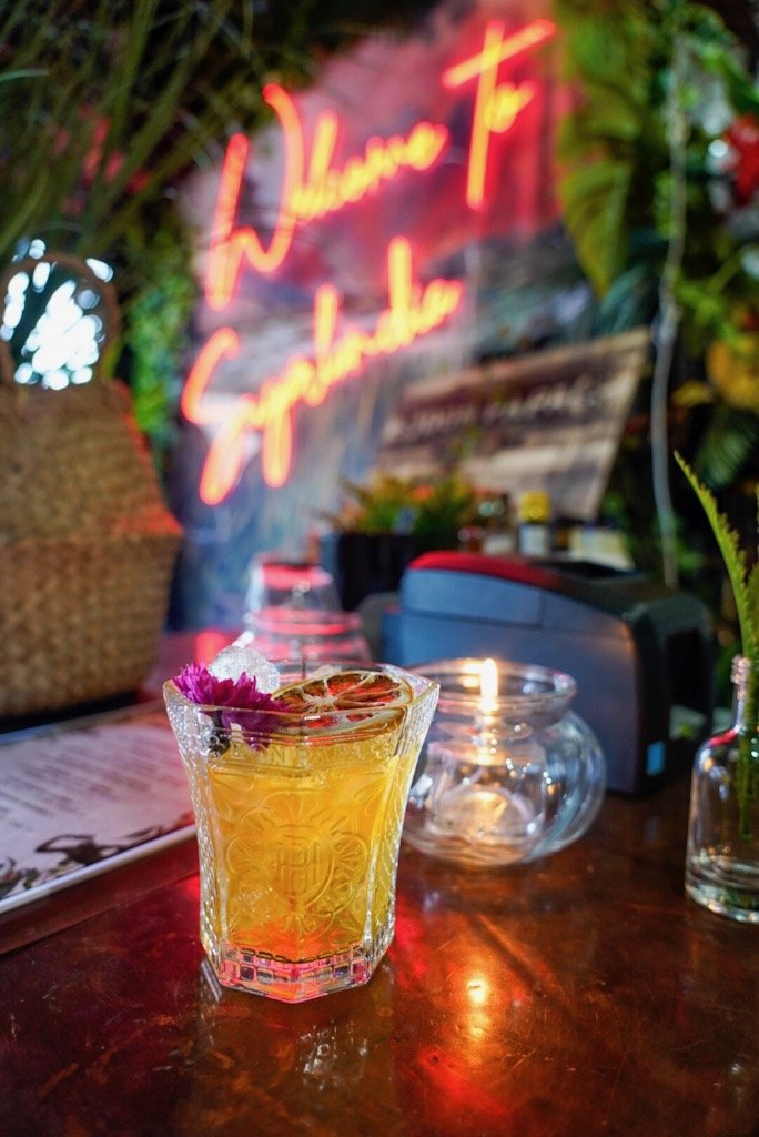 A cocktail garnished with a flower and citrus slice on a table, with a candle nearby and a neon sign in the background.