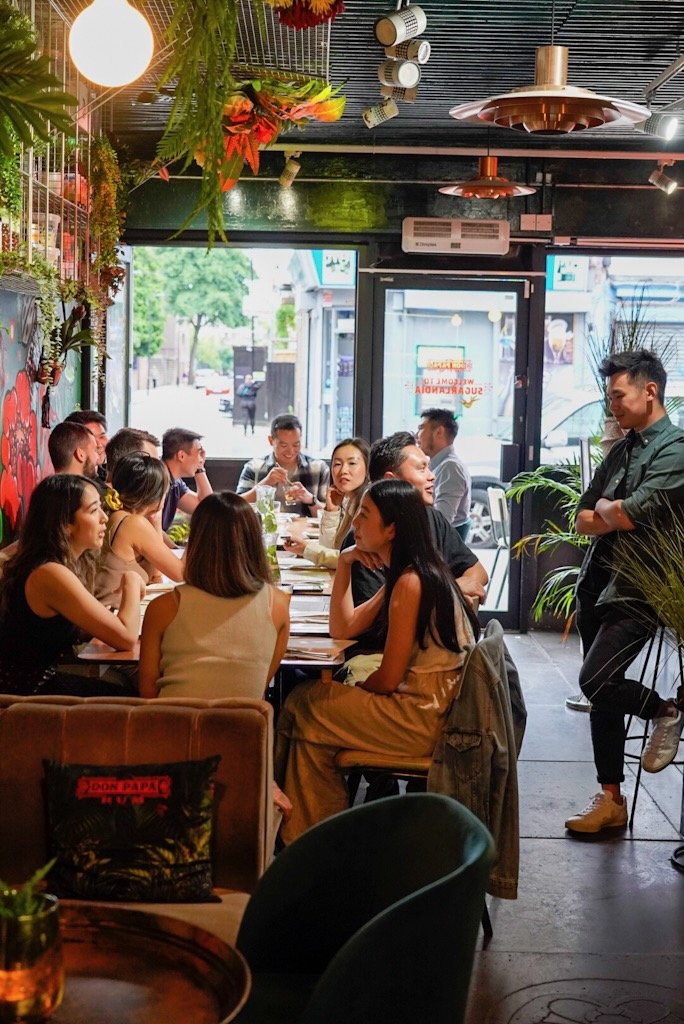 A group of people dining in a modern restaurant with plants and hanging lights, engaged in conversation around a long table.
