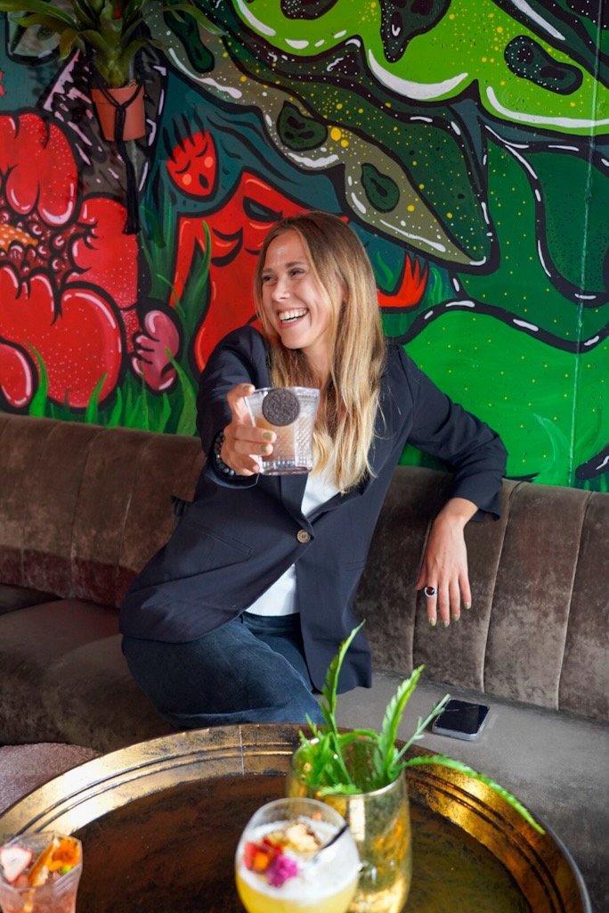 A woman in a black blazer holding a drink with a decorative cookie, sitting on a couch in front of a colorful graffiti wall with abstract and floral designs.