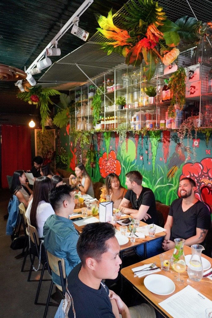 Group of people dining at a lively restaurant with colorful mural and hanging plants.