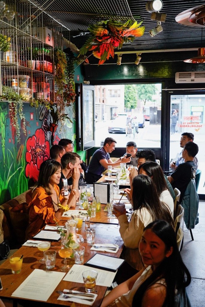 People sitting at a long table in a vibrant restaurant with colorful wall art, plants, and drinks on the table.