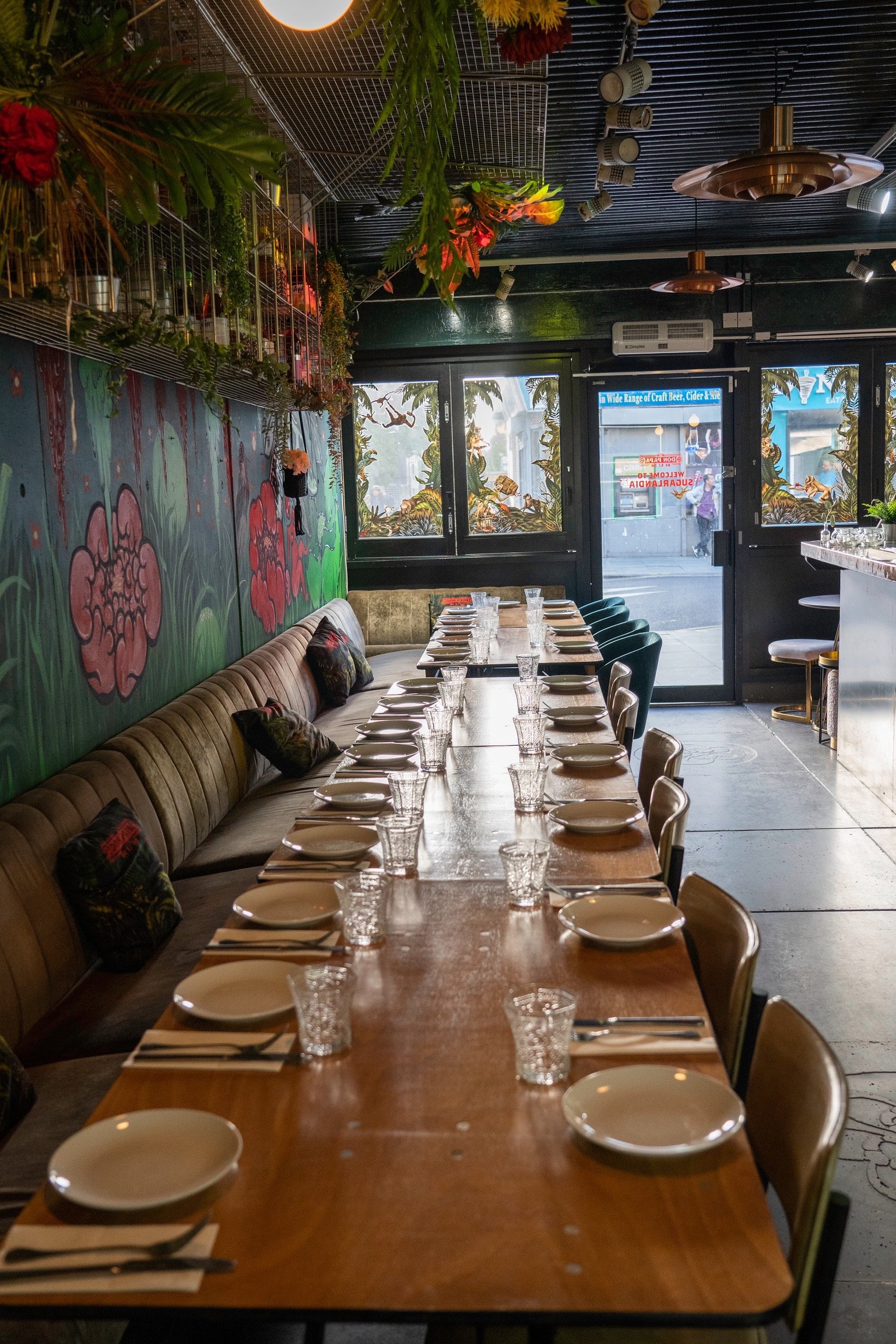 A long table in a restaurant set with plates and glasses, surrounded by chairs. The interior is decorated with vibrant floral and plant-themed murals and hanging greenery. The setting is colorful and inviting, with large windows providing natural lig