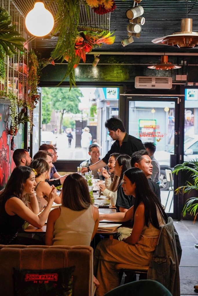Group of people dining at a long table in a restaurant with modern decor and hanging plants.