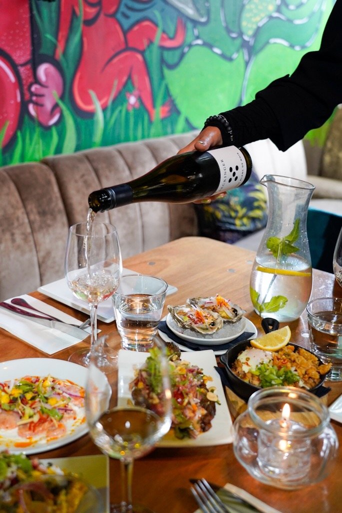Person pouring white wine into a glass on a table with various dishes including oysters, lettuce-topped salads, and a skillet dish. There's also a water carafe and a candle.