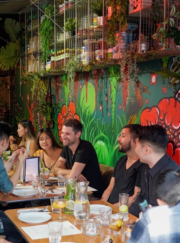 A group of people dining in a restaurant with colorful wall art and greenery. The table is set with glassware and plates. There are shelves with plants and various items above them.