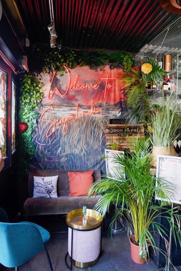 Colorful, cozy lounge area with a "Welcome to Sugarlandia" neon sign on the wall, surrounded by green plants and vibrant decor. A small sofa with decorative pillows sits next to a round table. Tropical-themed aesthetics dominate the space.