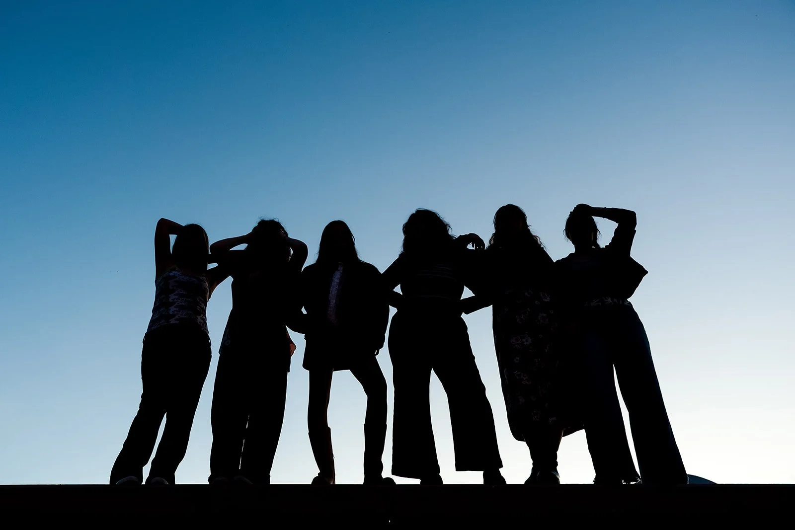 Silhouettes of six women standing together against a clear sky at sunset.