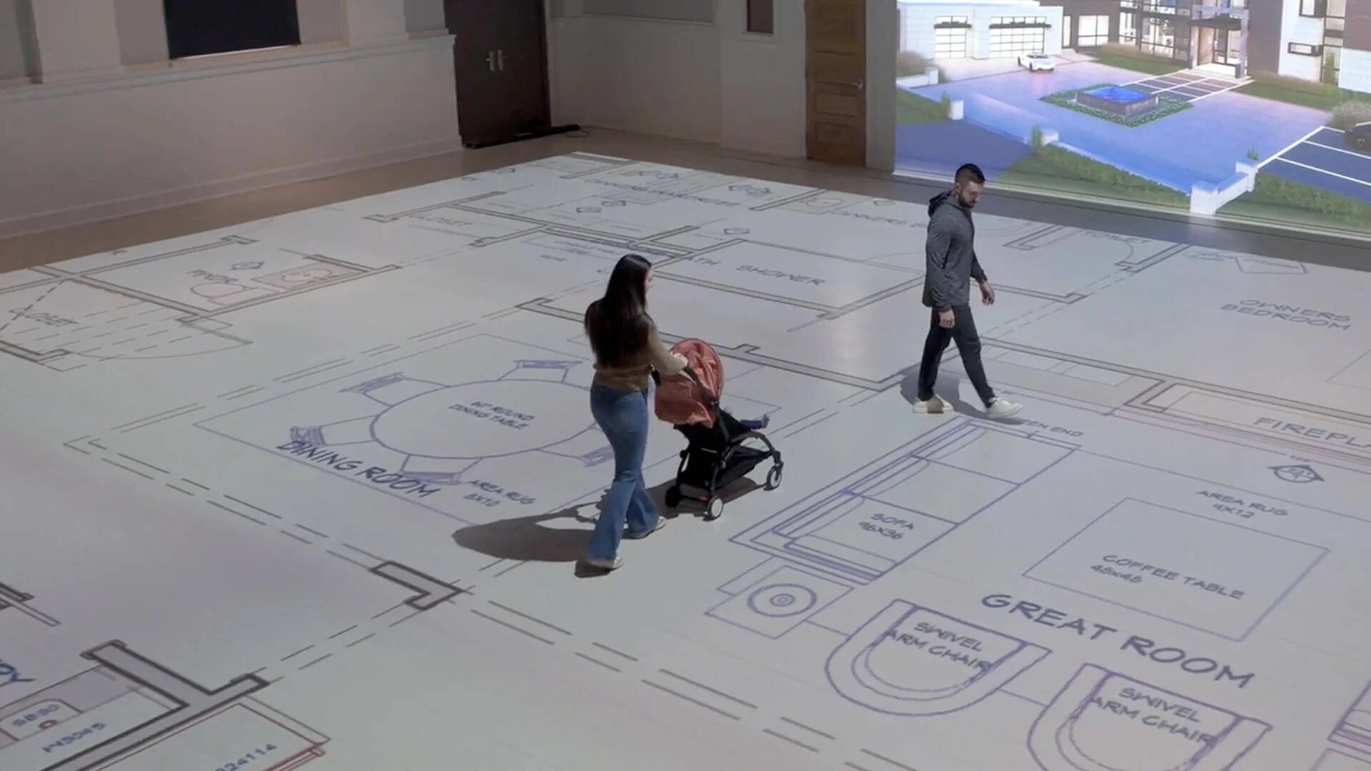 A woman pushing a stroller and a man walking inside a room with a projected floor plan of a house, including labels for rooms like living room, sitting area, great room, kitchen, and outdoor patio.