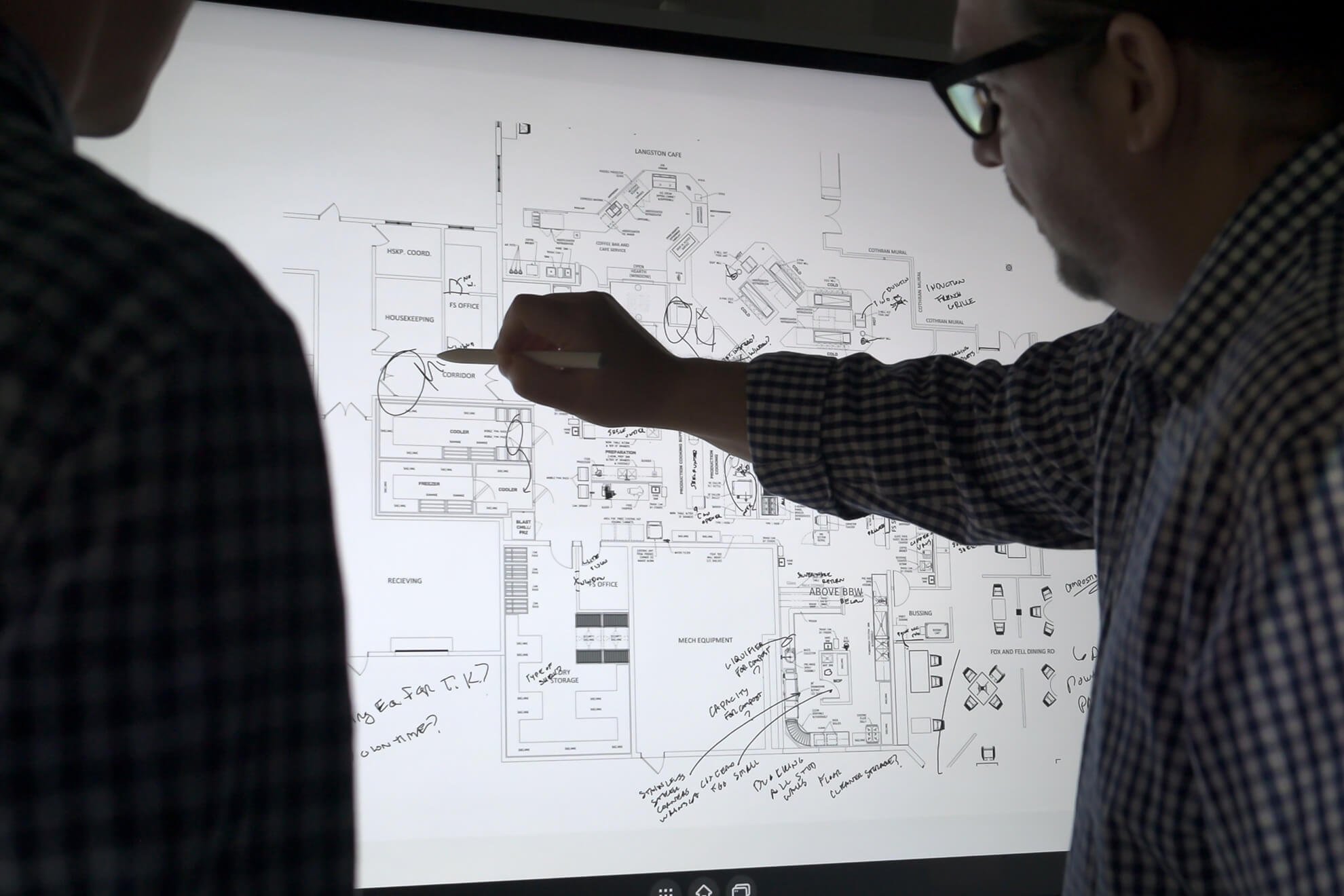 Two people examining a large architectural floor plan with handwritten notes, drawn on an illuminated lightbox.