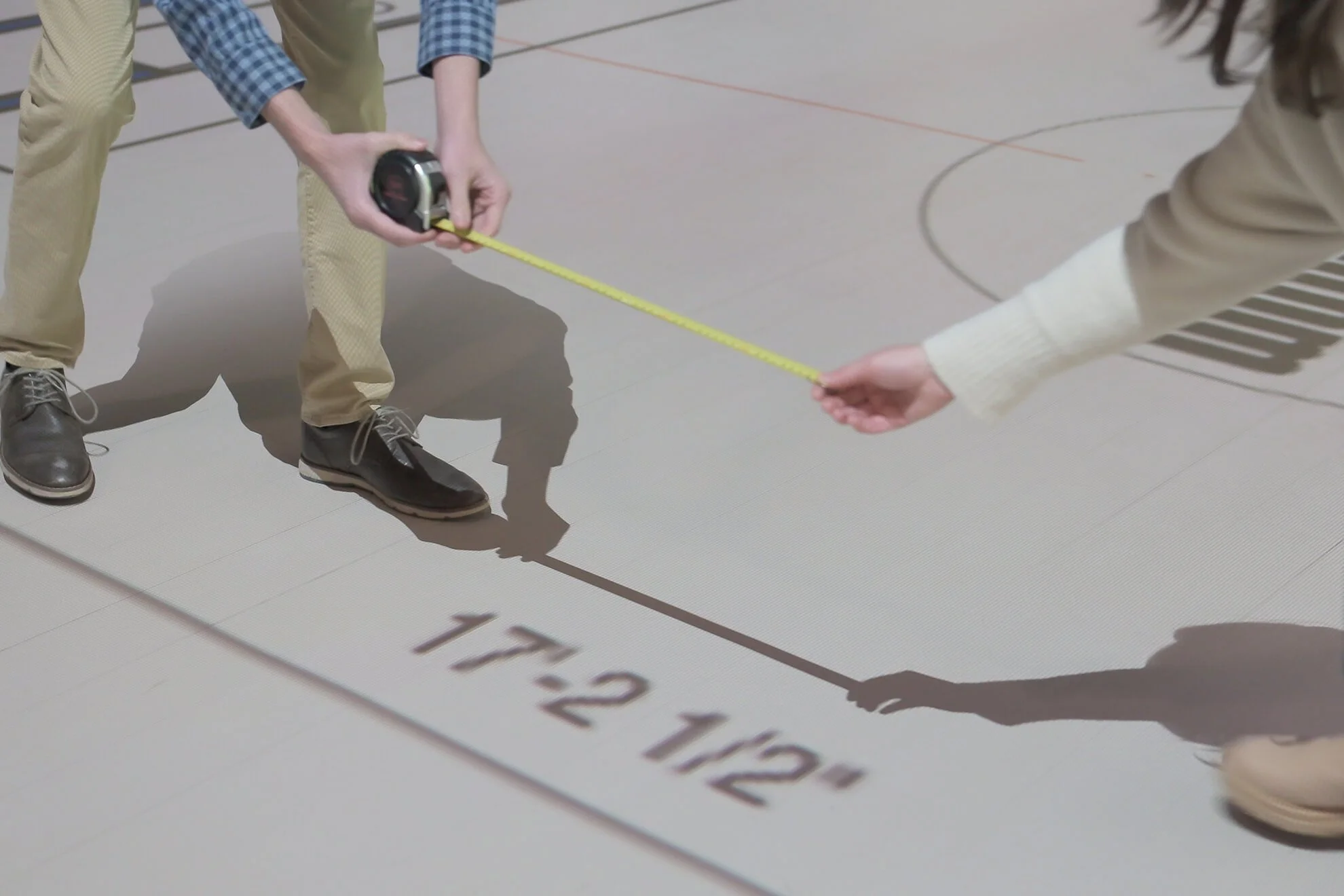 Two people measuring distance on the floor with a tape measure. One person is holding the tape measure, and the other is reading the length. The floor has markings and a label reading 'T 2 12'.