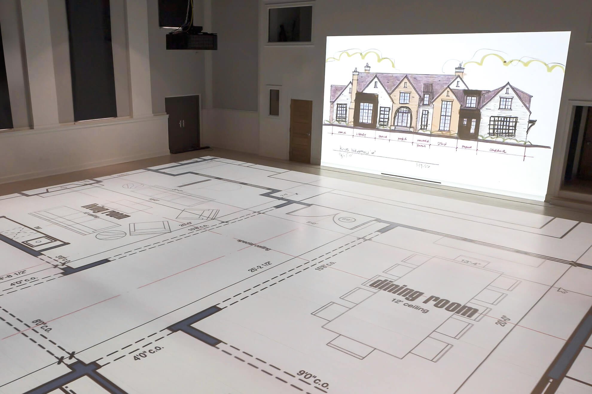 Architectural blueprint of a house layout projected on a floor in a room, with a drawing of a front house elevation on the wall.