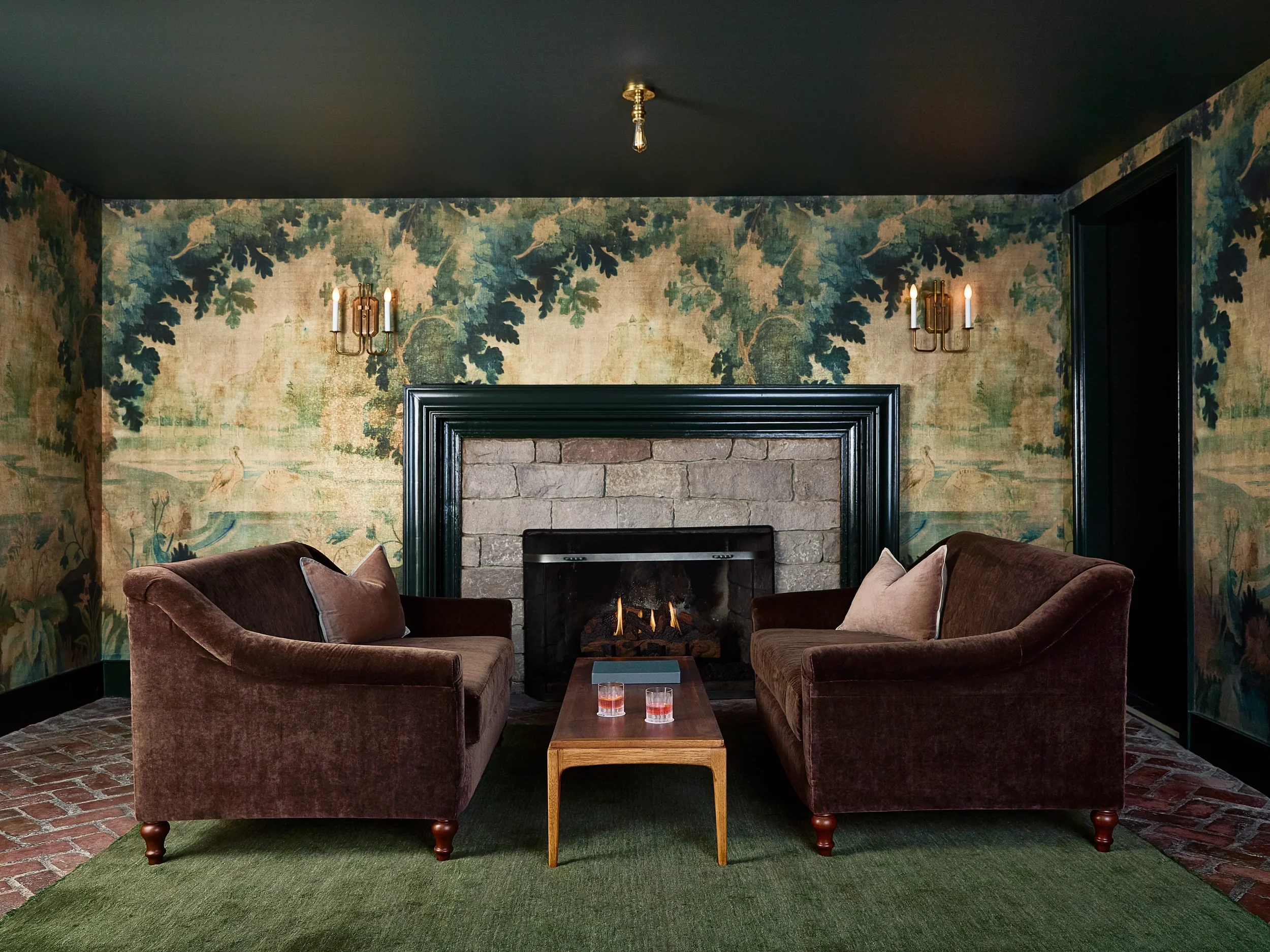 Living room with a stone fireplace, two brown velvet sofas facing each other, a small wooden coffee table with three glasses of water, floral wallpaper, brass wall sconces, black ceiling, and hardwood floor with a green rug.