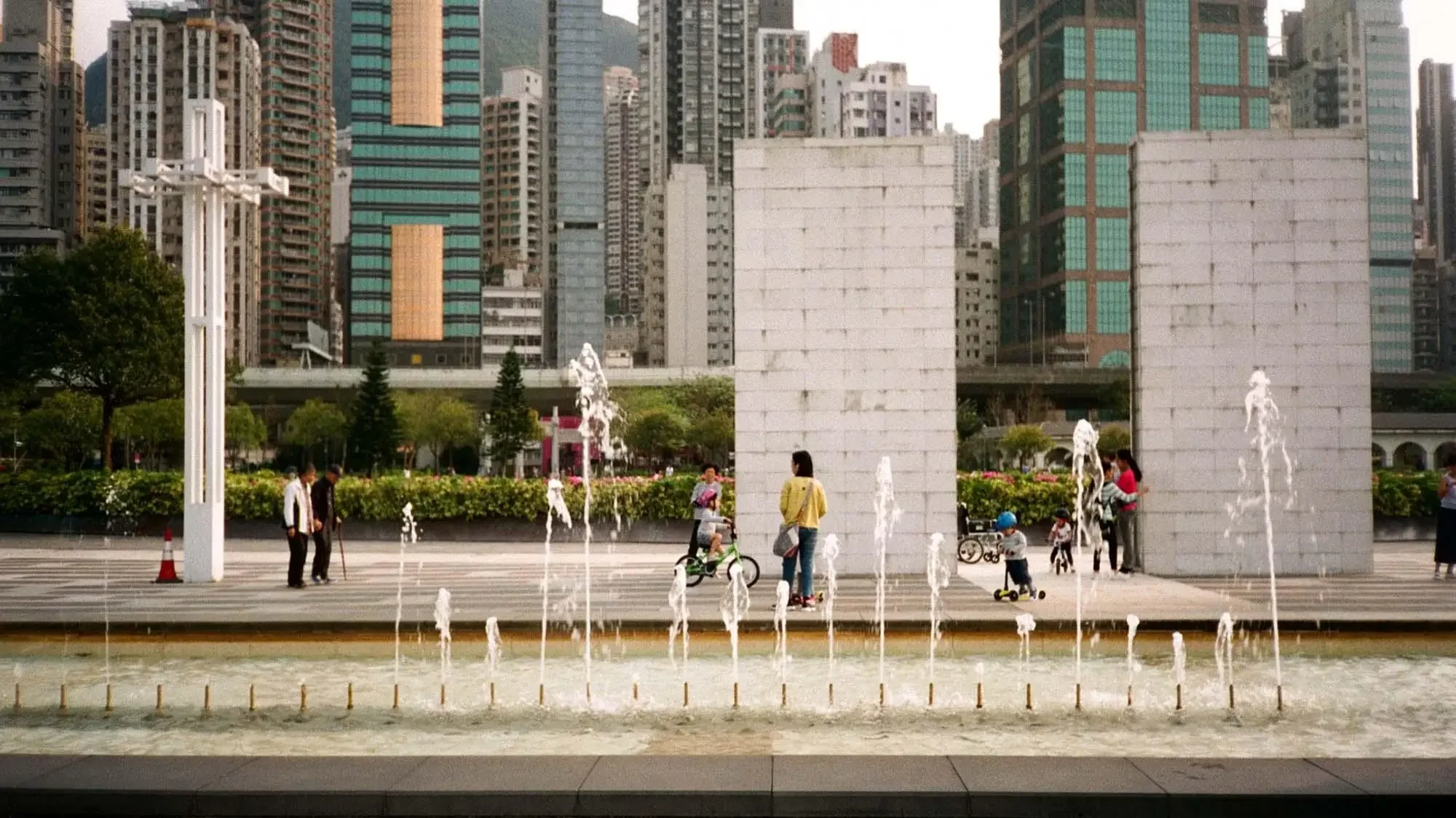 People in a city park with a fountain, modern skyscrapers in the background, children riding scooters, adults walking, and large concrete structures.