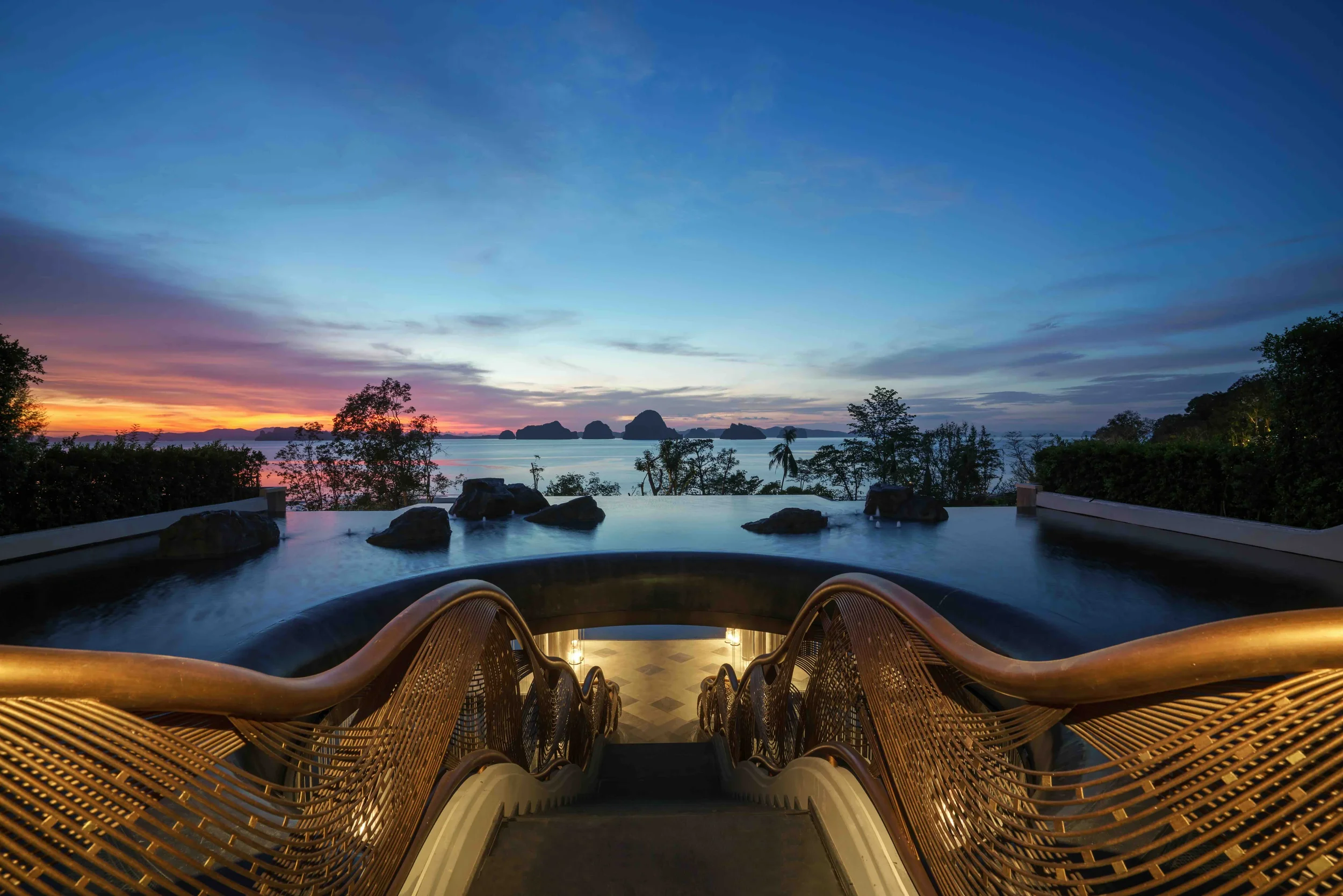 View from a staircase leading down to an infinity pool overlooking a sunset over the ocean with islands in the distance, surrounded by trees and bushes.