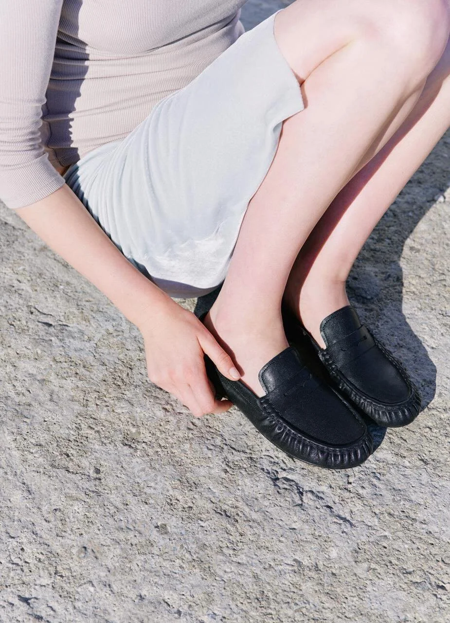 A person sitting on a rocky surface adjusting black slip-on shoes on their feet. The individual is wearing a light-colored, long-sleeve top and a light skirt.