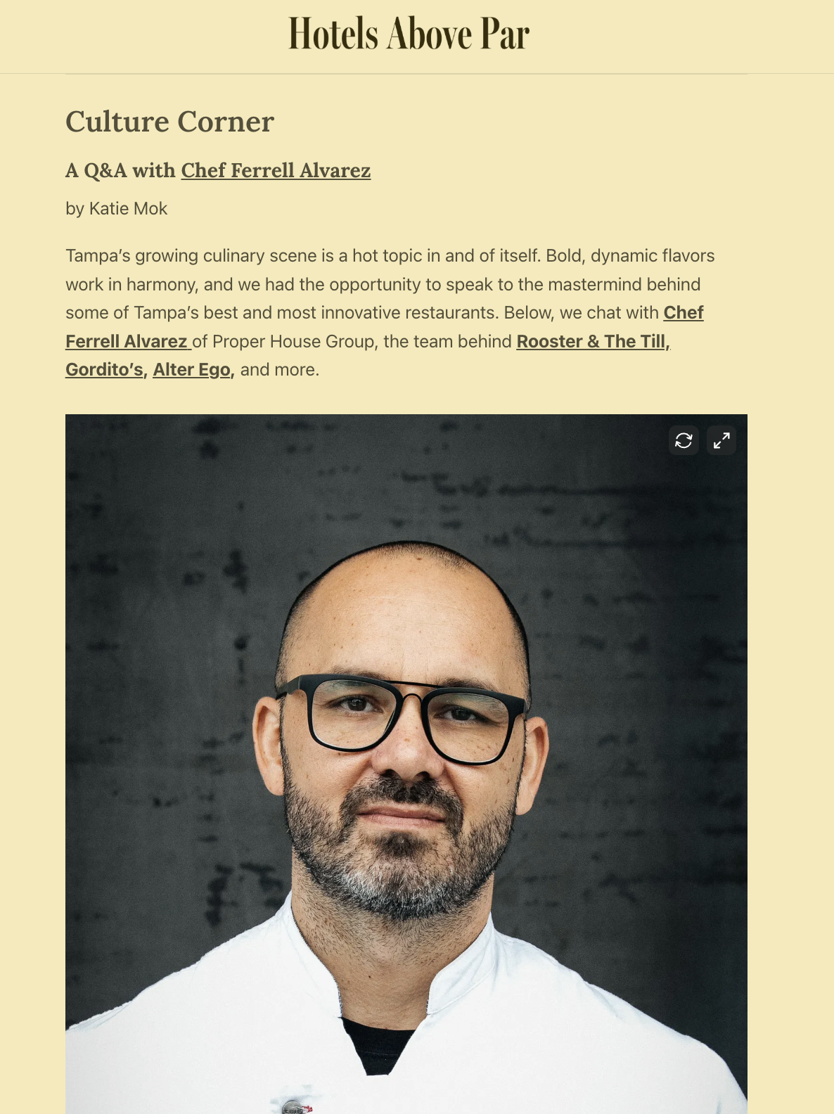 Portrait of a man with glasses, a beard, and a bald head, wearing a white chef's coat, standing in front of a dark textured background.