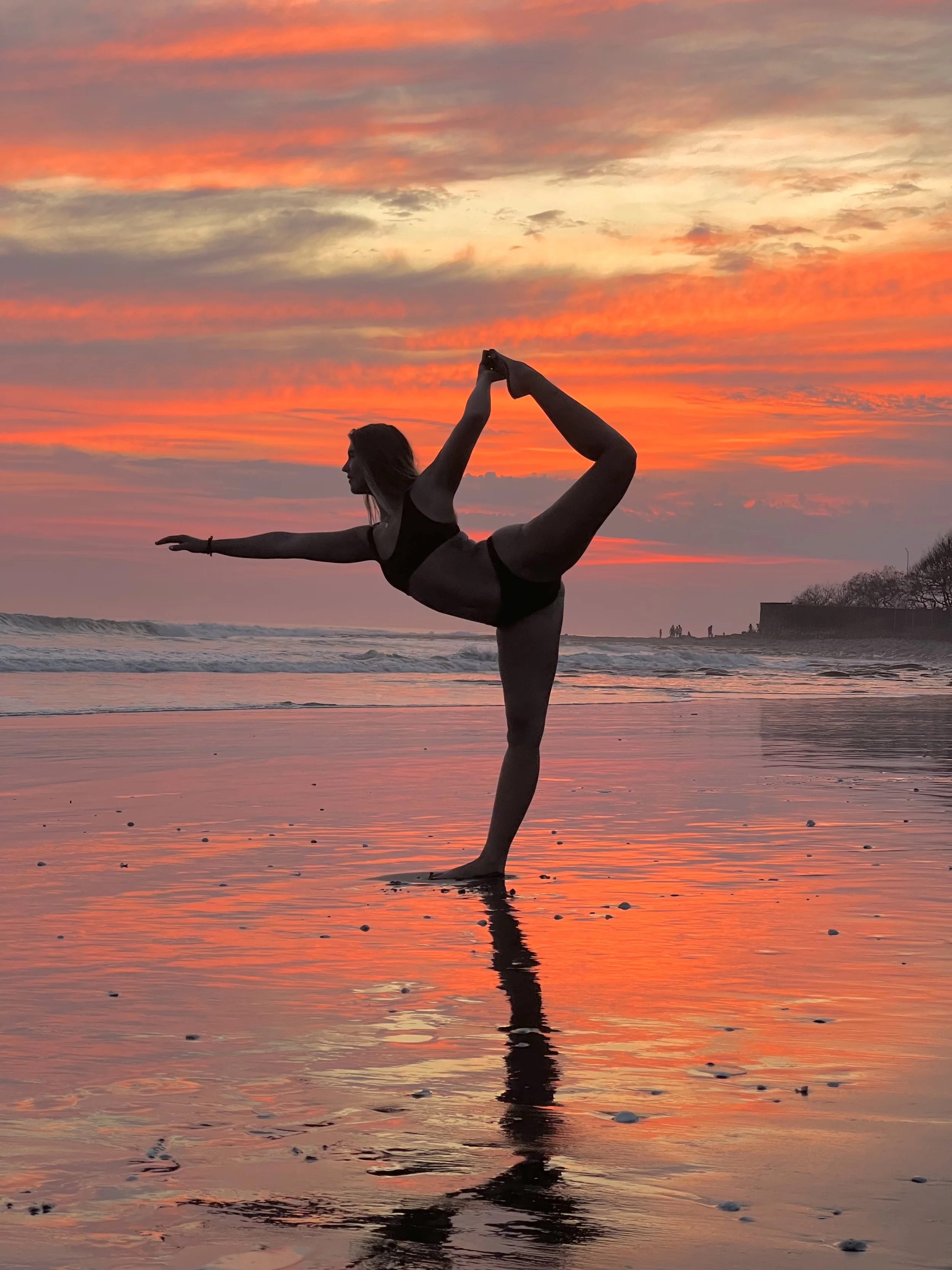 A woman practicing yoga in a dancer's pose on the beach at sunset, with colorful orange and pink clouds reflected in the wet sand.