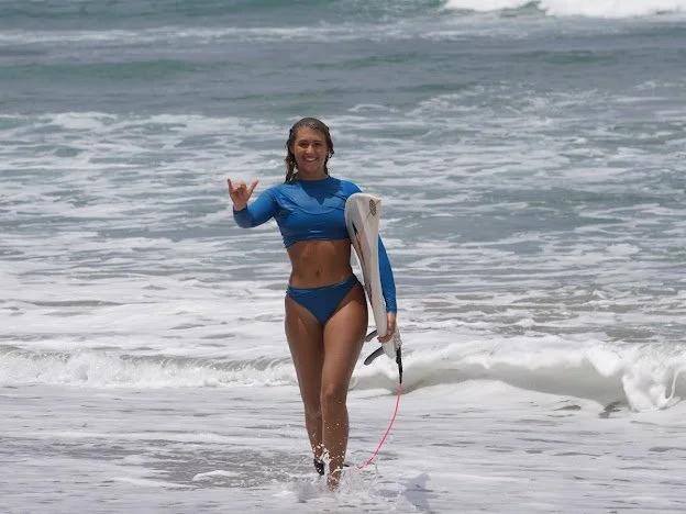 A woman in a blue swimsuit and matching long-sleeve top holding a surfboard, standing in the ocean waves near the shore, making a shaka sign with her right hand and smiling.