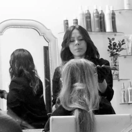 A woman getting her hair styled at a salon, seen in a mirror, with hair products in the background.