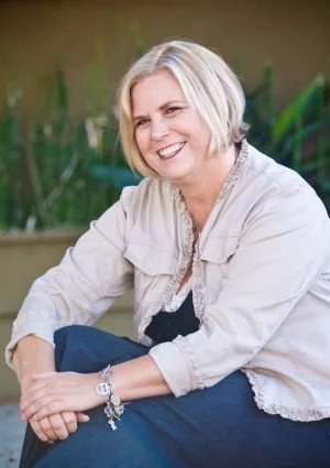 A woman with short gray hair, smiling, sitting outdoors with green plants in the background.
