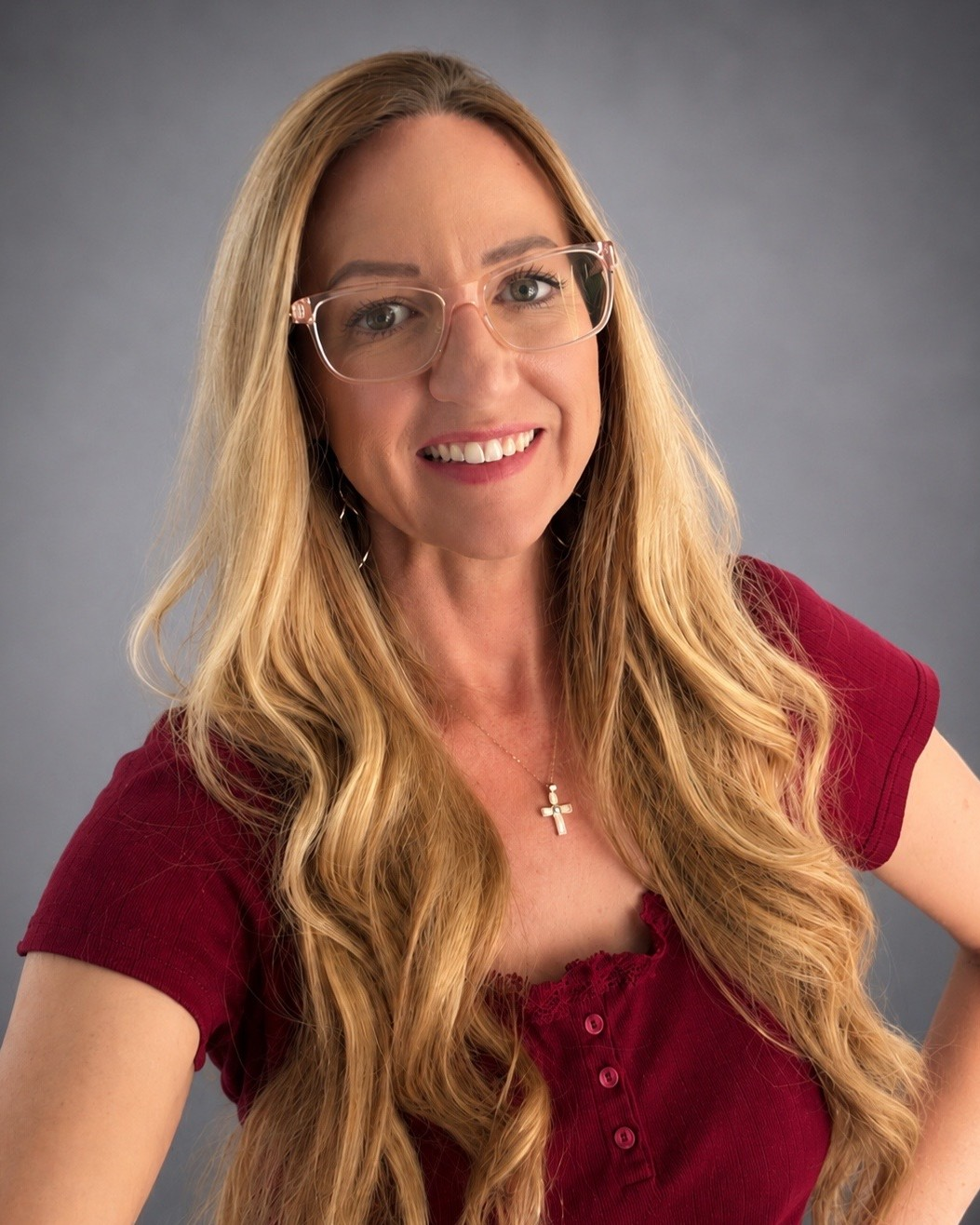 A woman with glasses and earrings smiling for the camera, with a light-colored textured wall in the background.