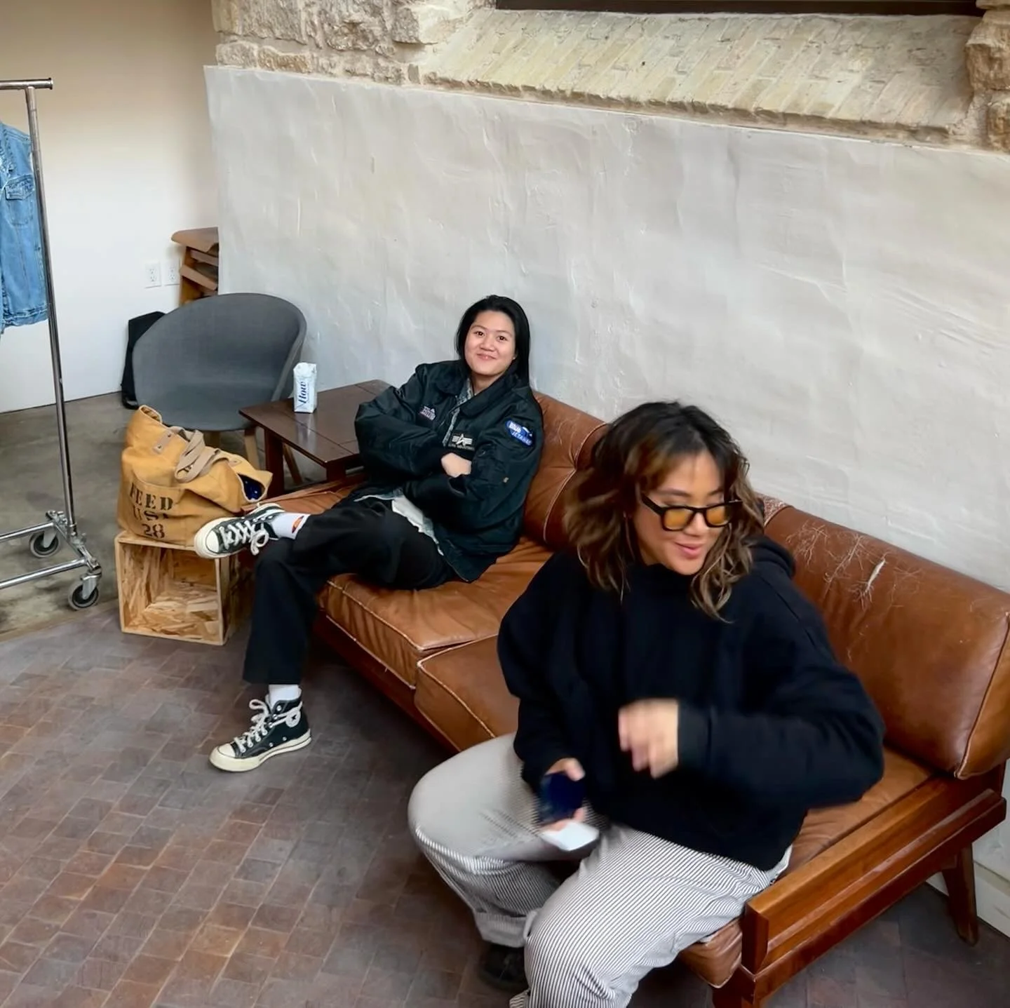 Two people sitting on a brown leather couch indoors. One person is wearing a black jacket and sneakers, smiling with arms crossed. The other person is wearing glasses and a black top, looking down. A clothing rack and a tote bag are in the background